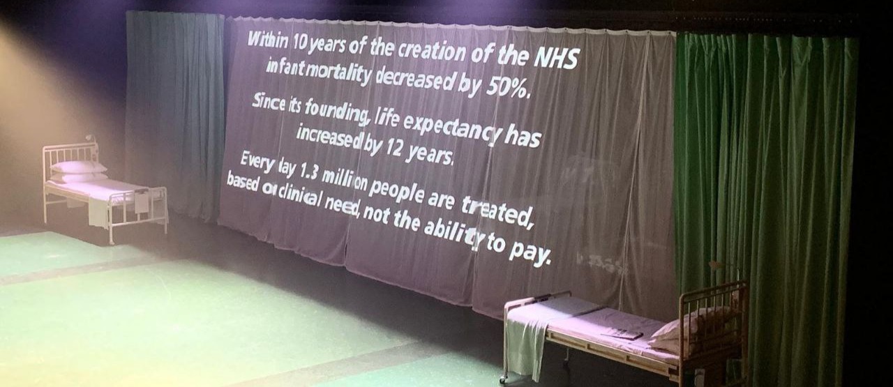 Curtain from production of ’Nye’, text: ’Within 10 years of the creation of the NHS, infant mortality decreased by 50%. Since its founding, life expectancy has increased by 23 years’.
